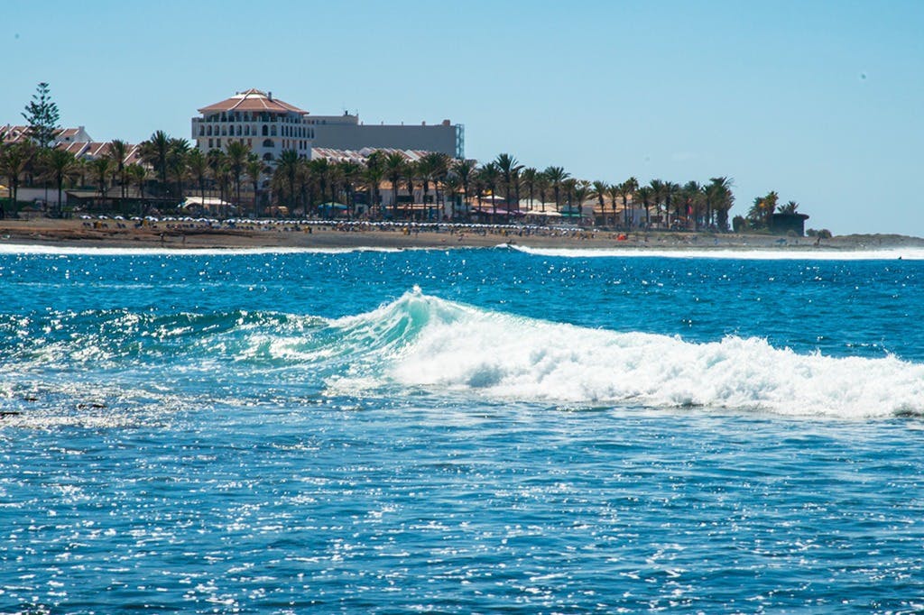 Волны катятся в океане с далеким видом на пляжный отель и линию пальм под ясным голубым небом.