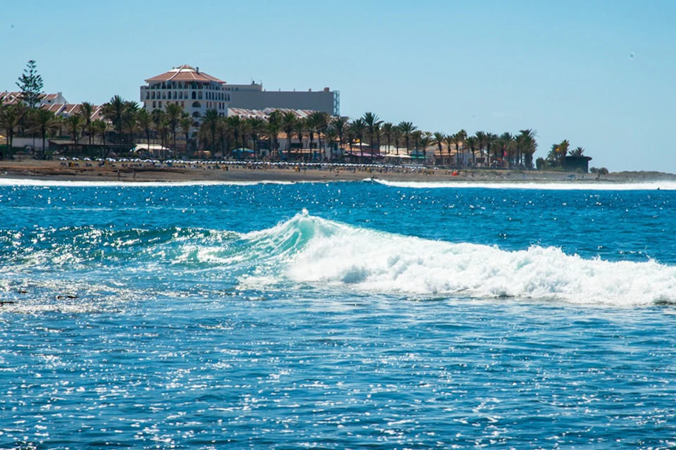Waves rolling in the ocean with a distant view of a beachfront hotel and a line of palm trees under a clear blue sky.