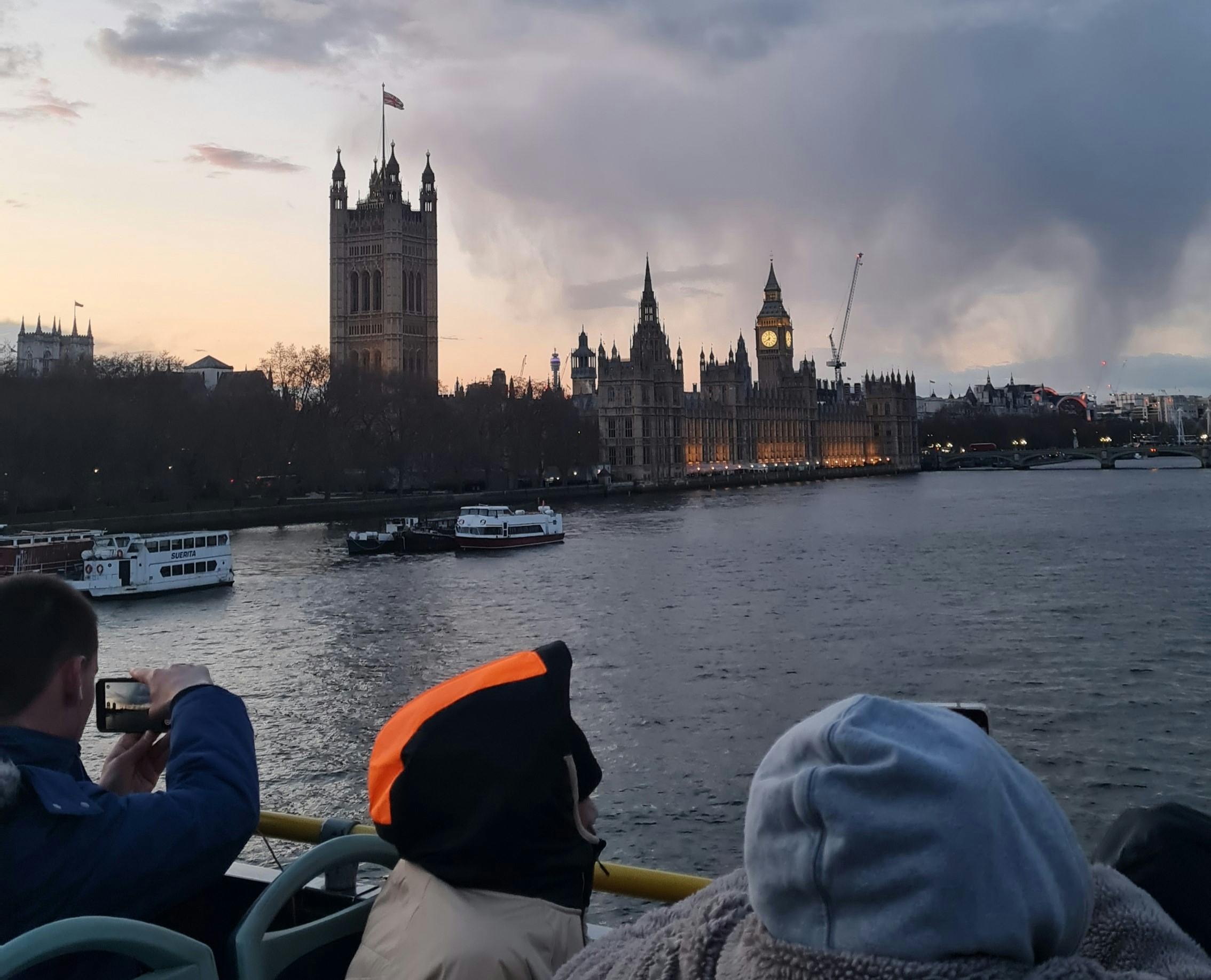 夕暮れ時、テムズ川を渡って照らされたビッグベンとウェストミンスター宮殿を眺めながら、暖かい服を着た人々がボートに乗っています。