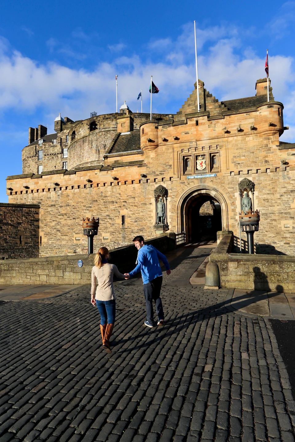 A man and woman walk hand-in-hand toward a large historic stone castle with flags and statues above the entrance.