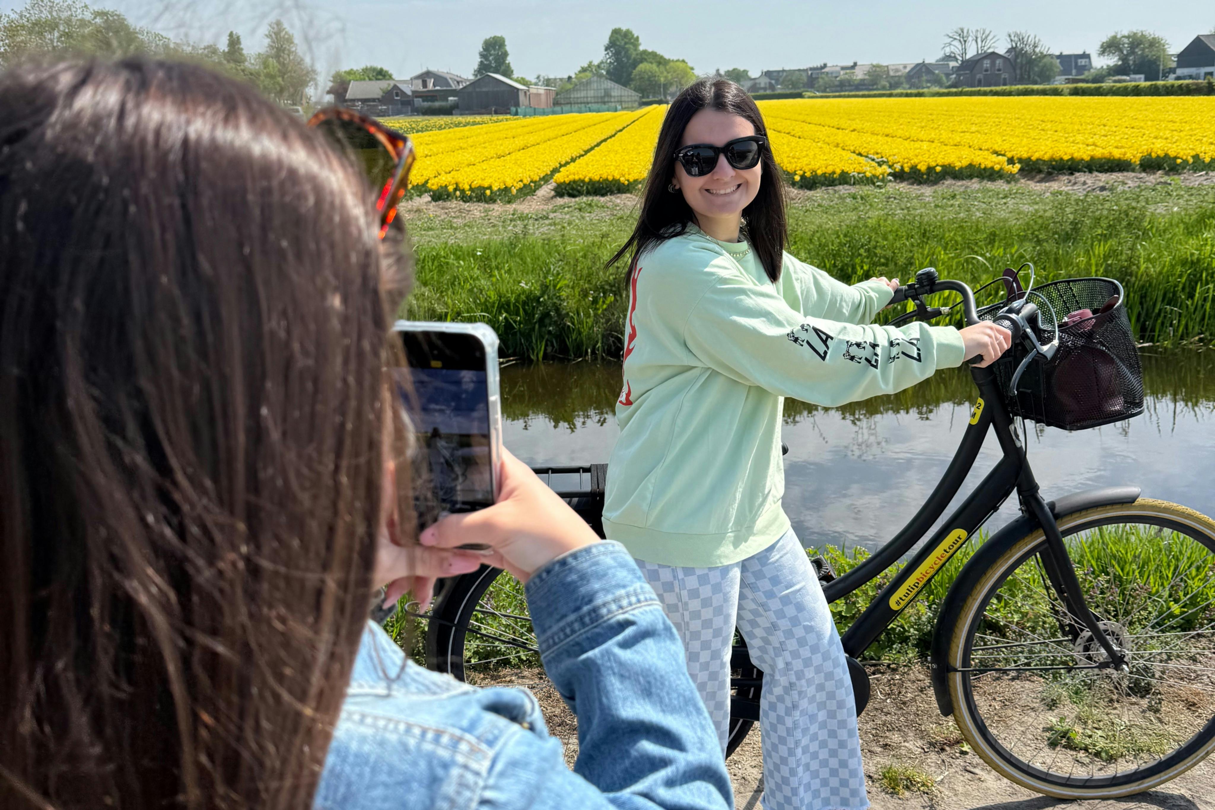 A woman on a bicycle, smiling at the camera, with yellow flower fields in the background. Another person is photographing her.