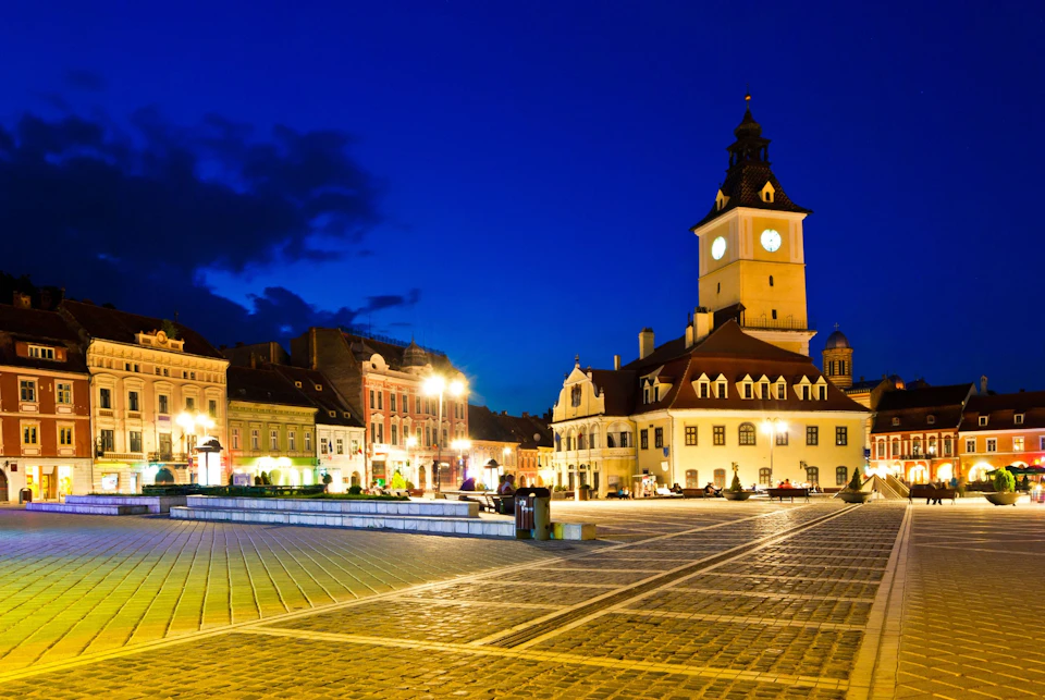 Town square at night with illuminated buildings, clock tower, and empty cobblestone plaza under a deep blue sky.