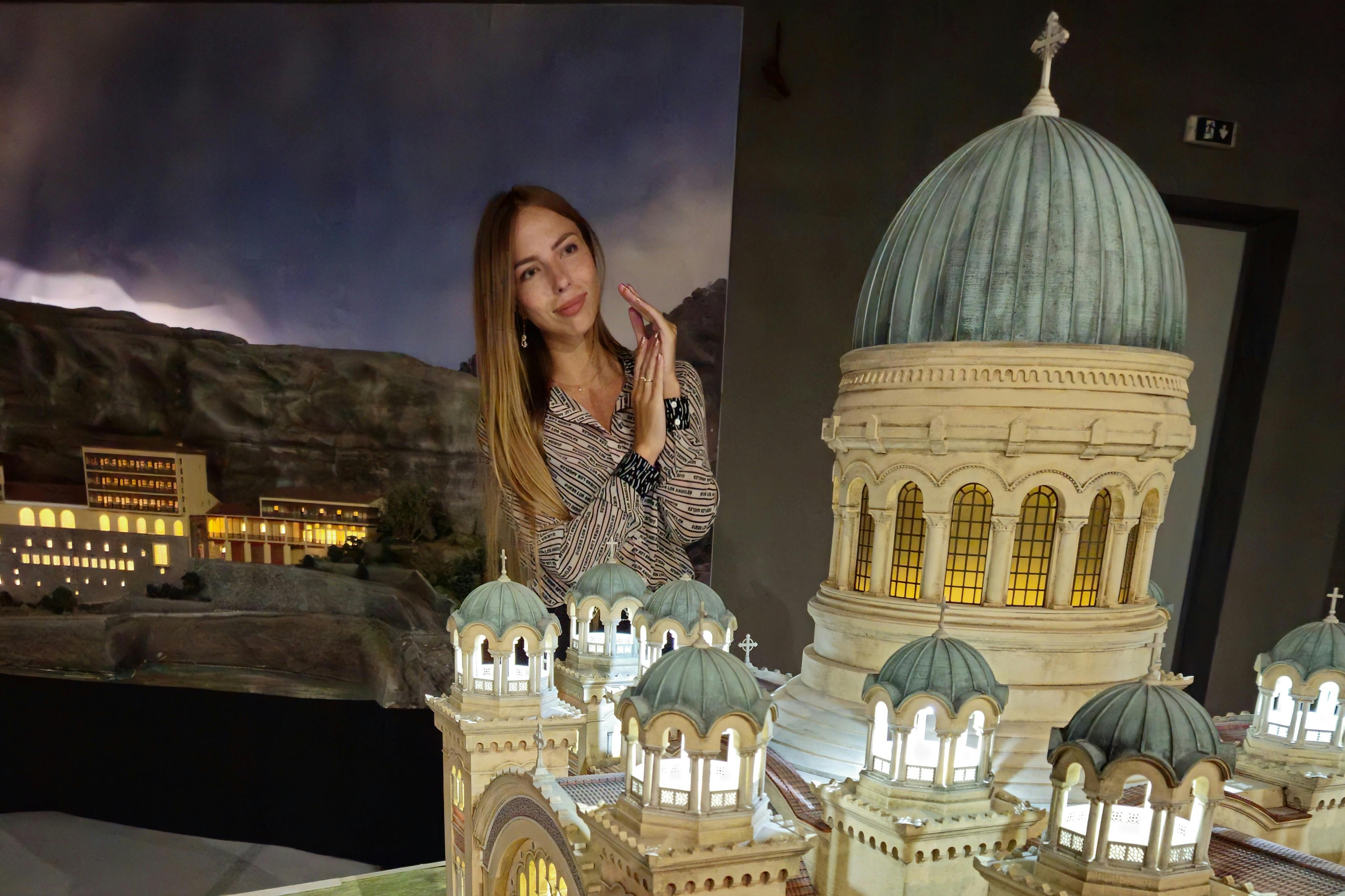 A woman stands smiling in front of a detailed architectural model with domed towers and lit windows, against a backdrop of rocky terrain.