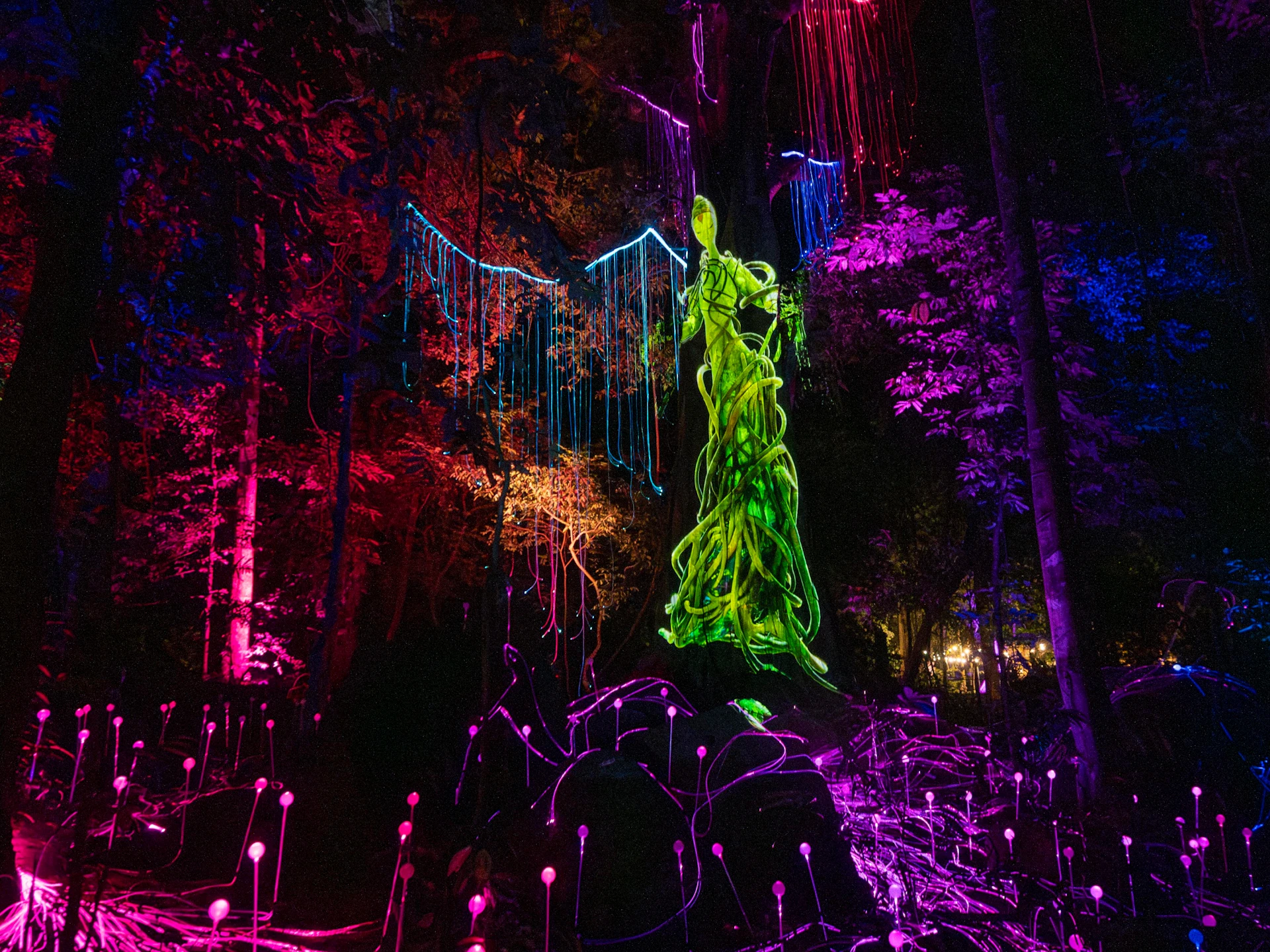 A glowing green statue surrounded by vibrant multicolored lights and illuminated trees in a nighttime forest setting.