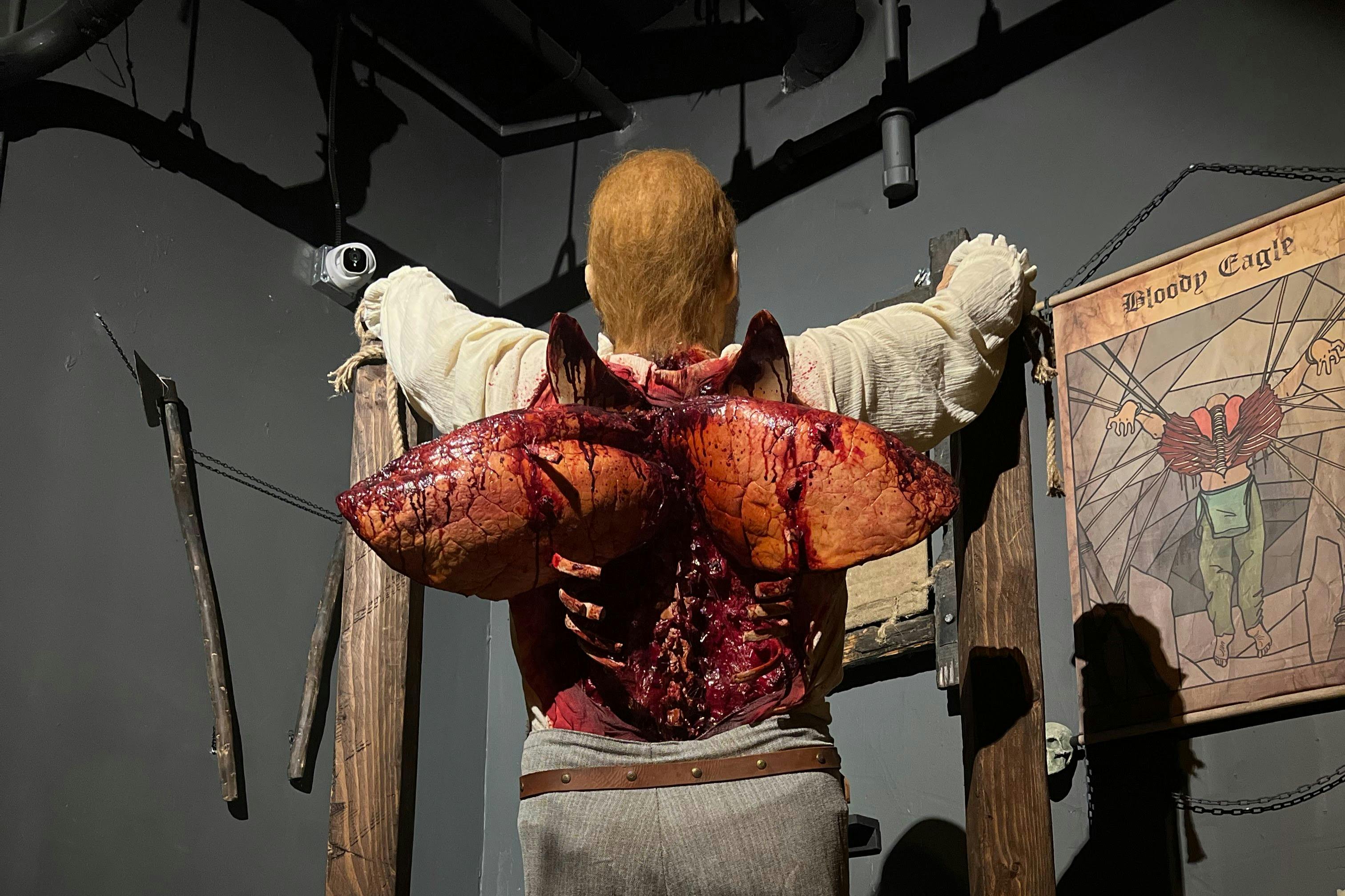 Ένα μανεκέν που απεικονίζει ένα άτομο με μια φρικιαστική, αιματηρή ανοιχτή πλάτη, τοποθετημένη σε ένα ξύλινο πλαίσιο σε ένα αμυδρά φωτισμένο δωμάτιο.