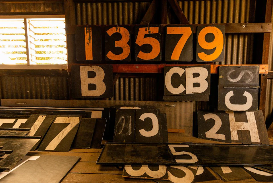 Various blackboards with orange and white numbers and letters are stacked against a corrugated metal wall in a dimly lit room.