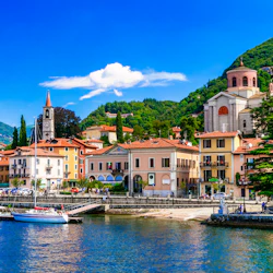 Färgglada byggnader och en kyrka vid en sjö, uppbackad av gröna kullar under en klarblå himmel.