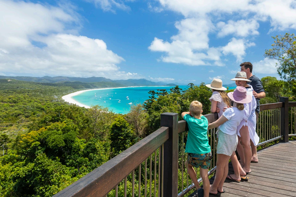 Four people stand on a wooden lookout, gazing at a tropical beach with turquoise water, boats, and a backdrop of lush greenery.