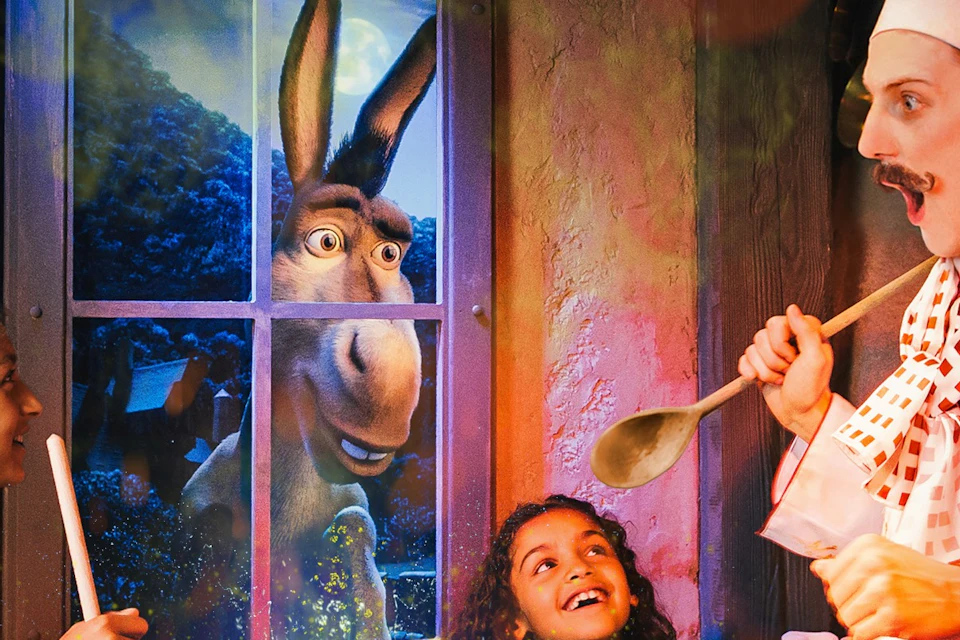 Two children and a chef stir a cauldron with green liquid, while a donkey looks in through the window, smiling.