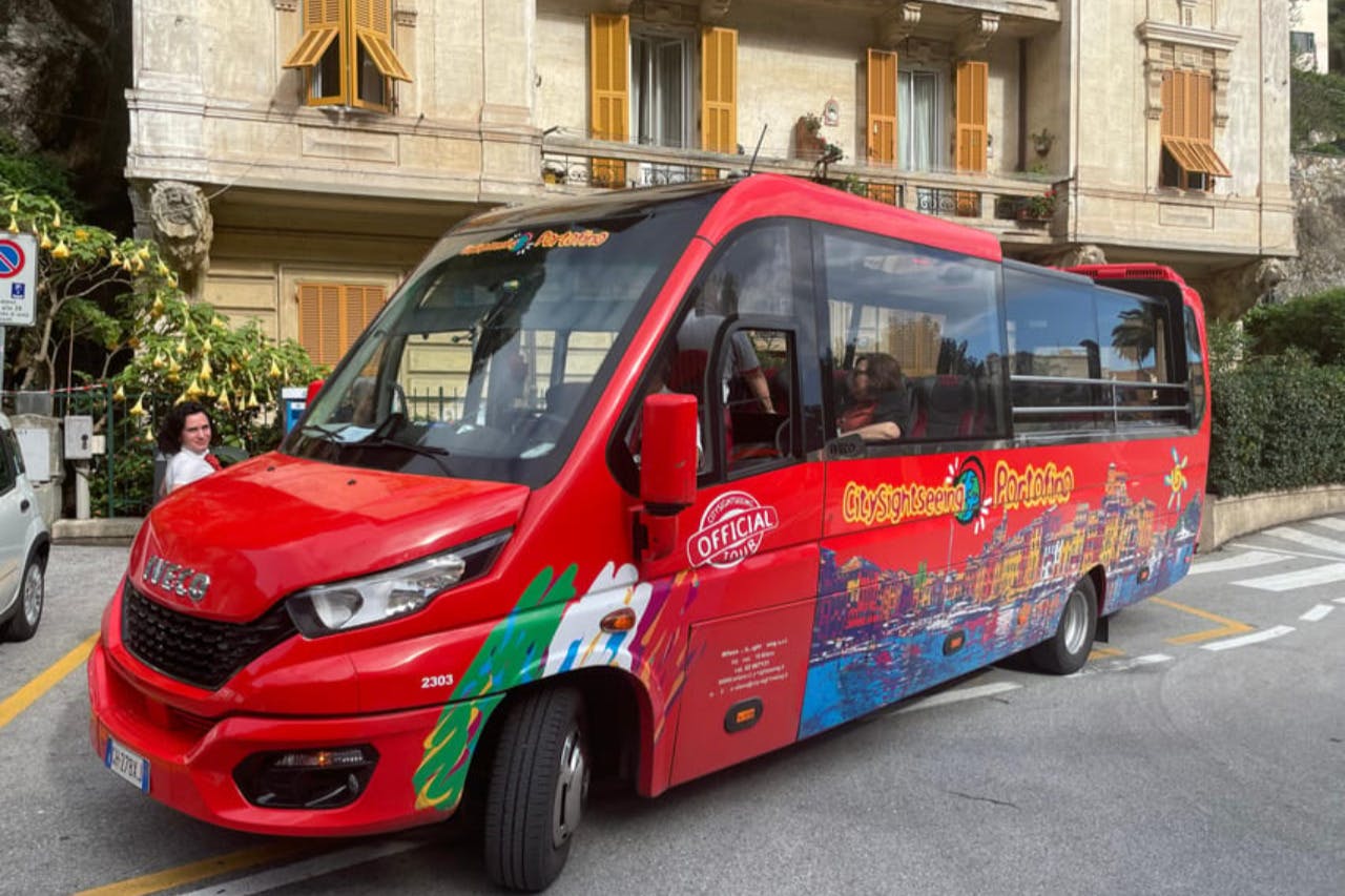 A red tour bus with colorful designs is parked on a street next to a beige building with yellow shutters and balcony plants.