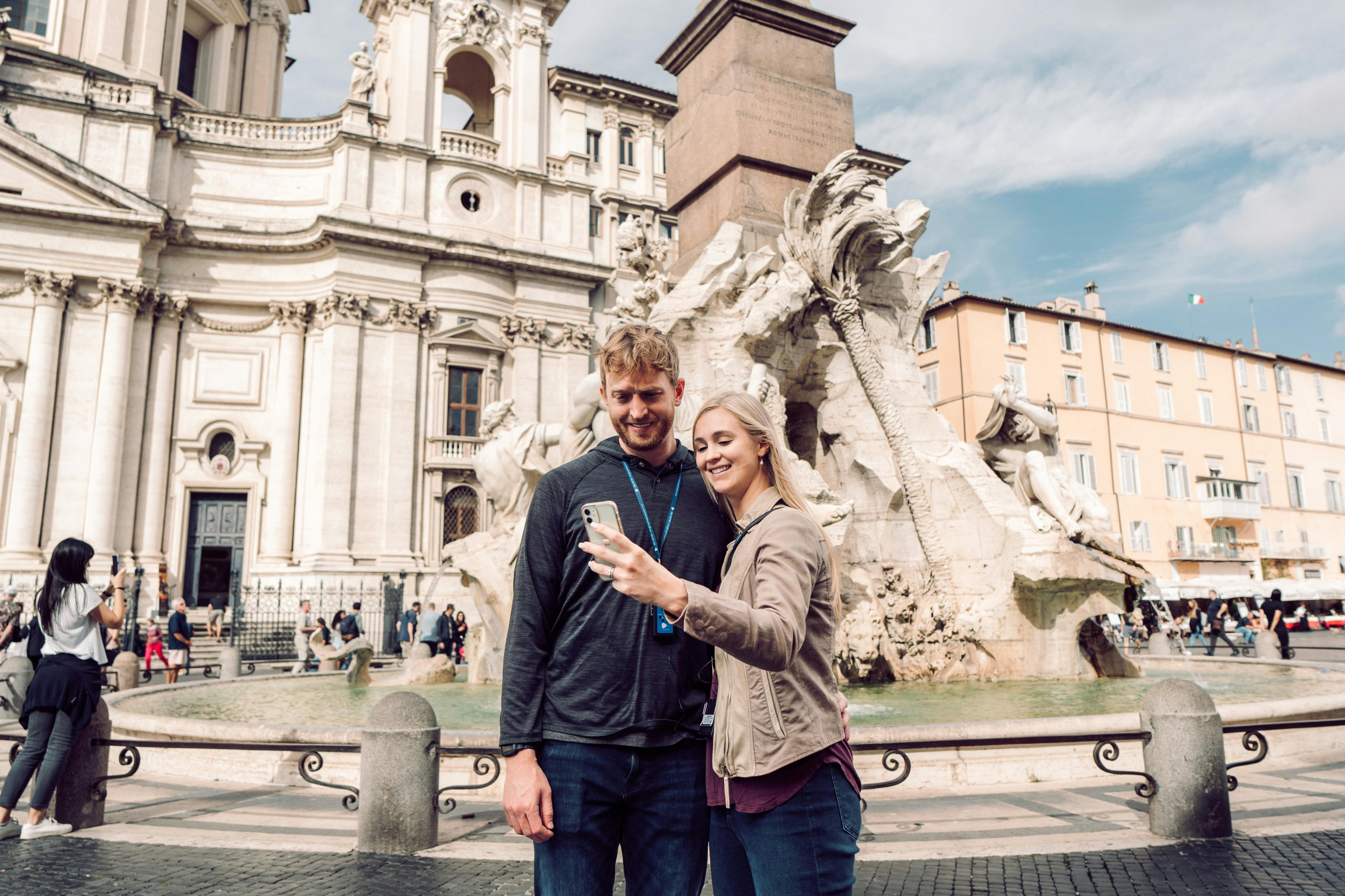 Una coppia sorridente si scatta un selfie davanti a una fontana storica e a un edificio decorato dall'architettura classica.