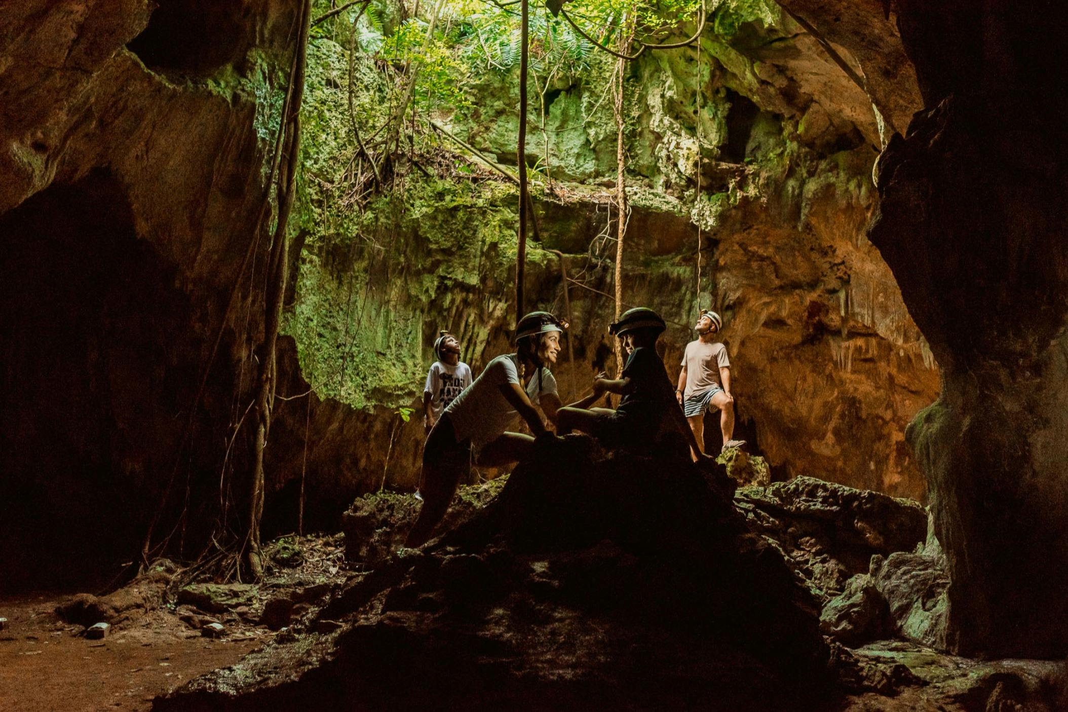 Four people exploring a lush, green cave with sunlight filtering through an opening above.