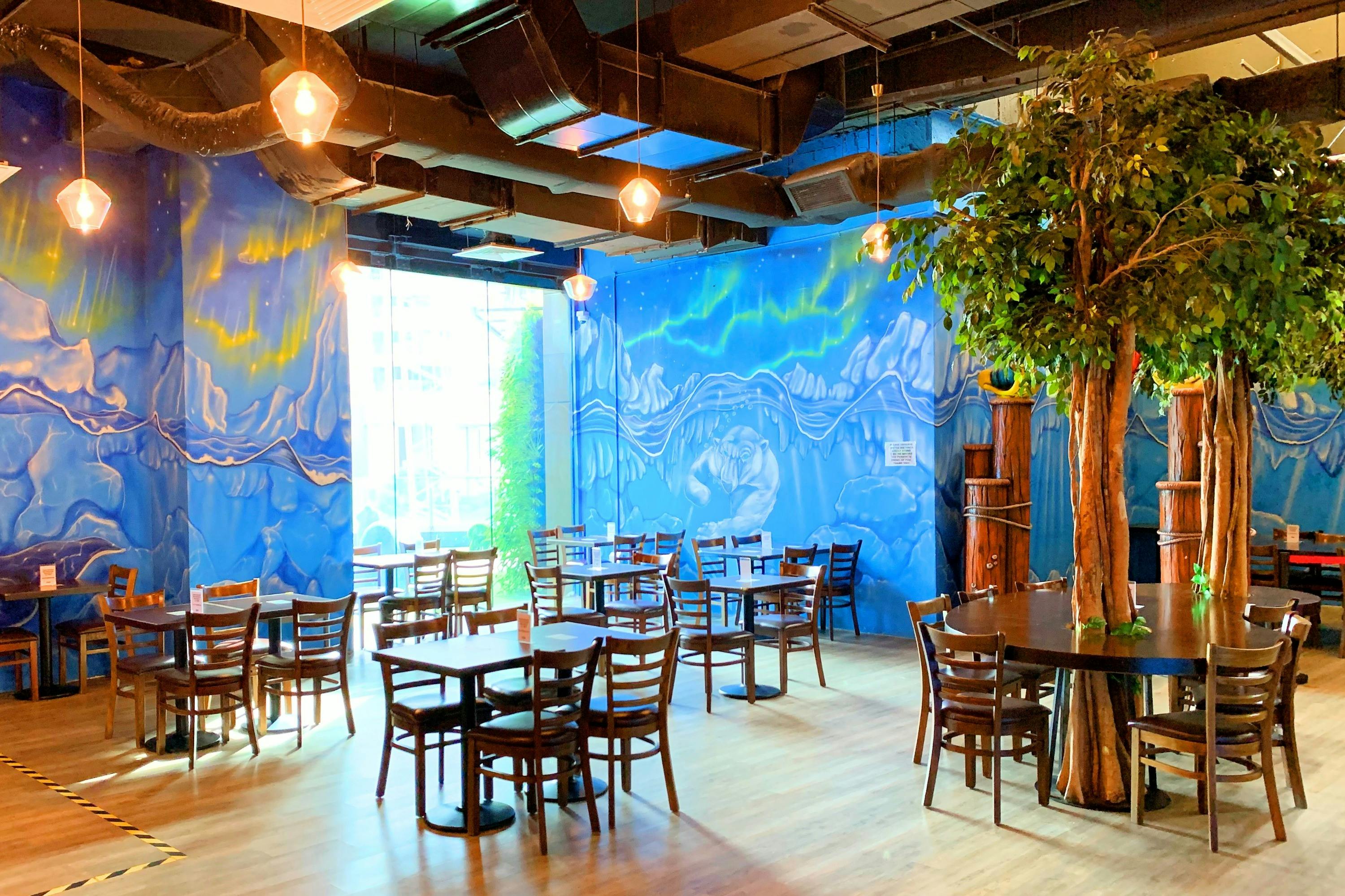 A brightly lit cafe with wooden tables and chairs, a blue mural on the walls, and decorative indoor plants.