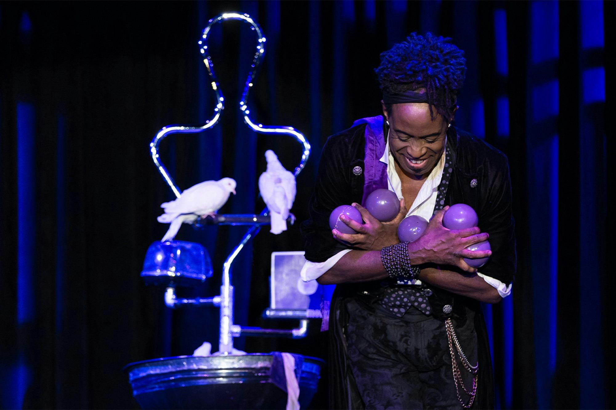 A performer on stage holding several purple balls while smiling, with doves and a lighted prop in the background.