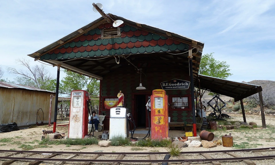 Old gas station with three vintage gasoline pumps, a wooden building with a B.F. Goodrich sign, and railway tracks in front.