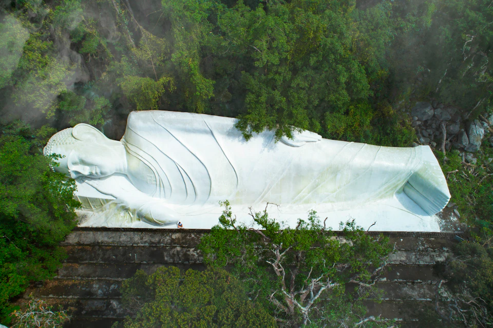 A large white reclining Buddha statue surrounded by dense greenery, with a small figure at its base for scale.