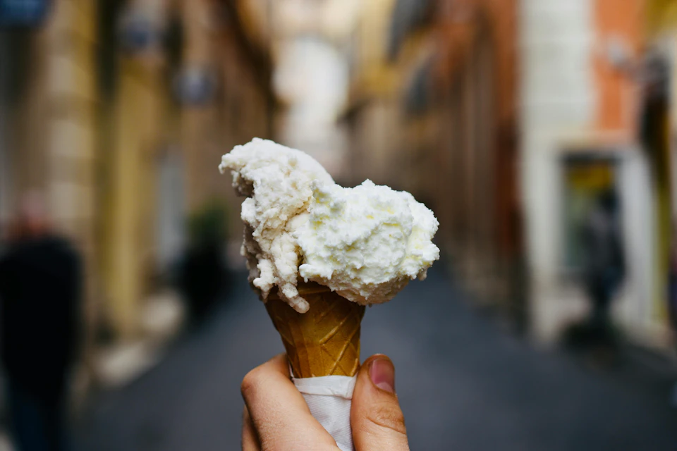 A hand holding an ice cream cone with two scoops of white ice cream in a narrow, blurred urban street.