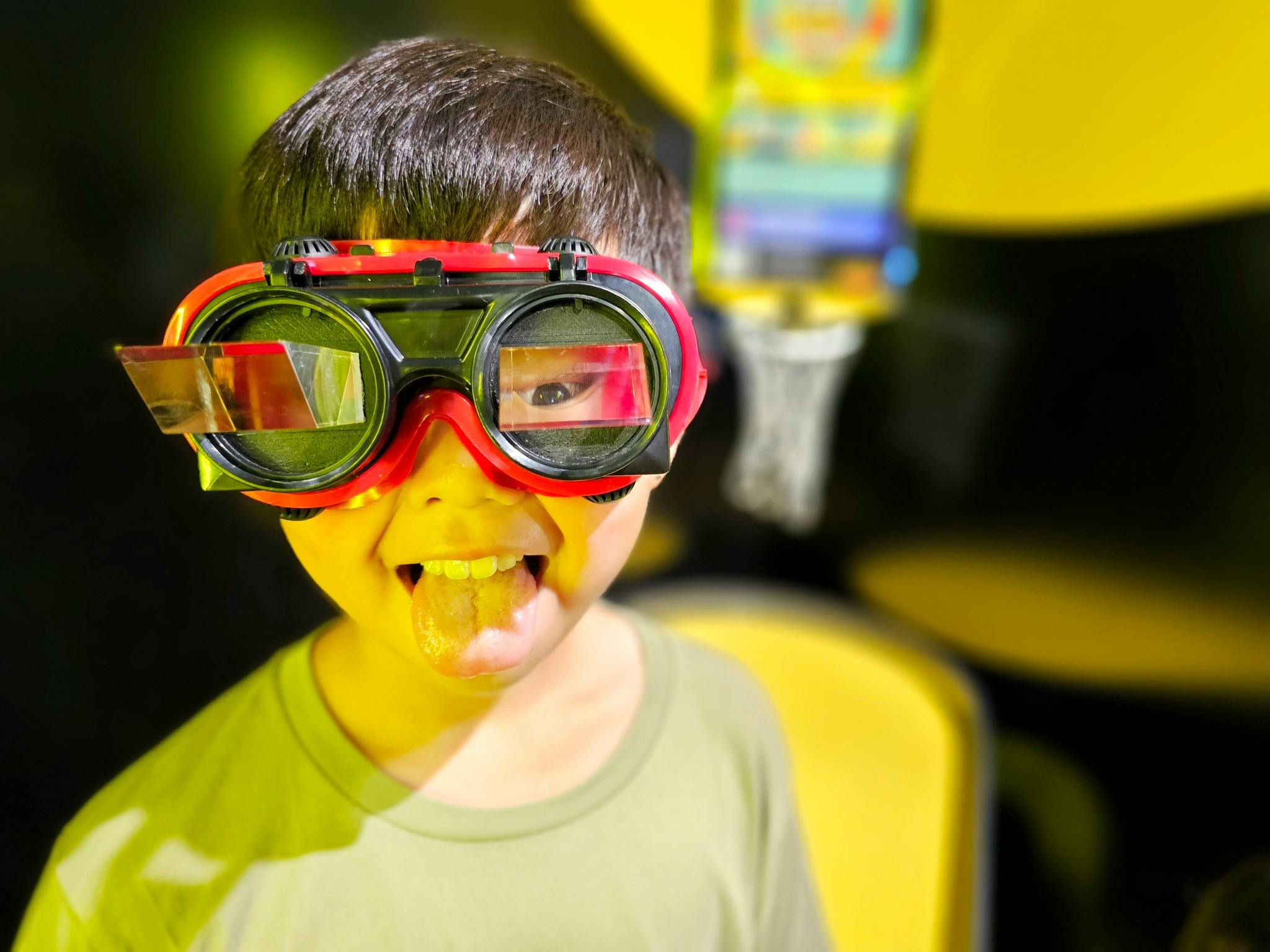 Child wearing oversized goggles with a playful expression, sticking out their tongue, in a brightly lit setting.