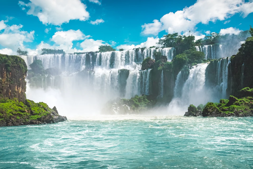 Waterfalls cascading over a rocky cliff into a wide river, surrounded by lush greenery under a blue sky with clouds.