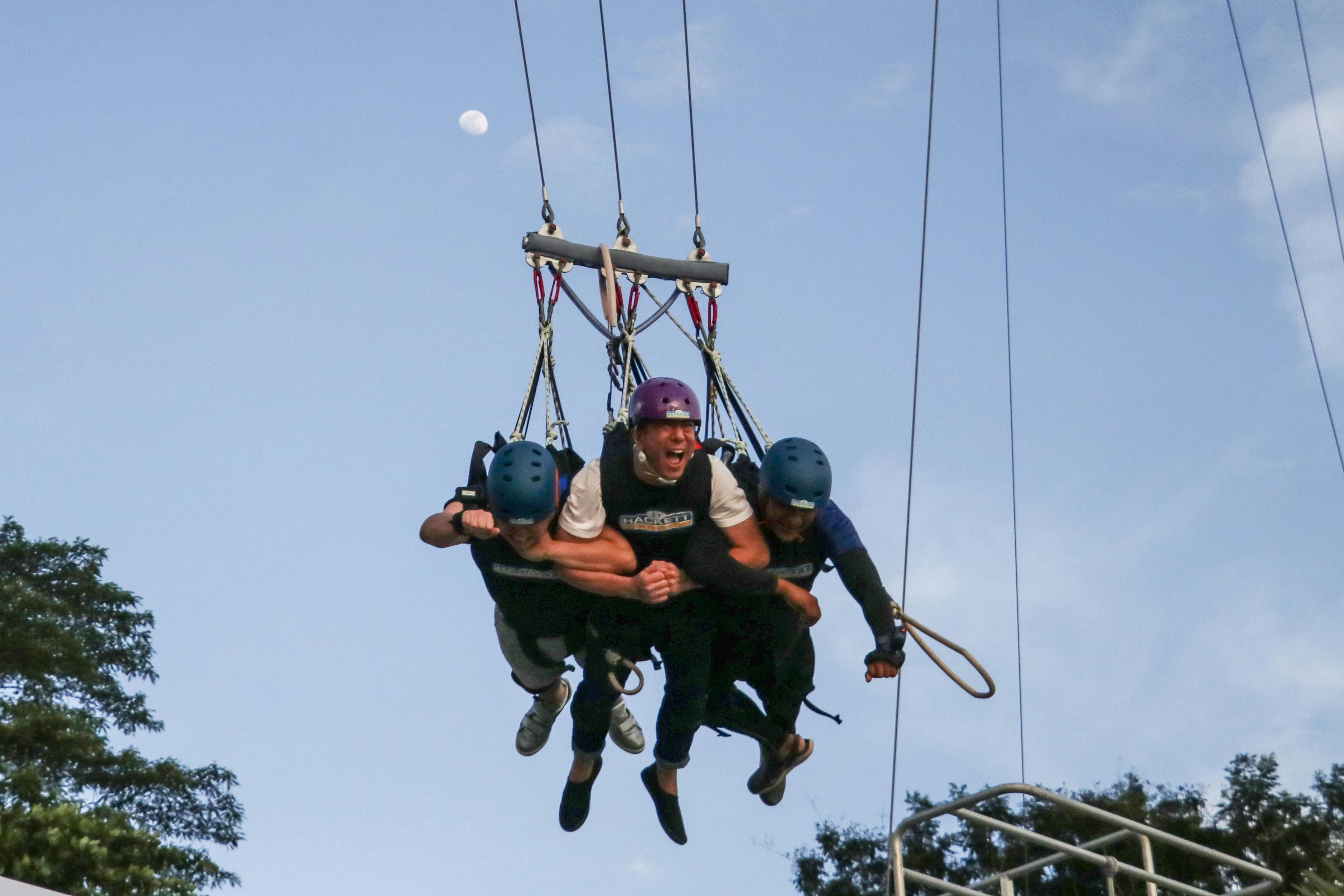 Three people with helmets are suspended and swinging in the air, holding onto each other tightly. The sky is clear.