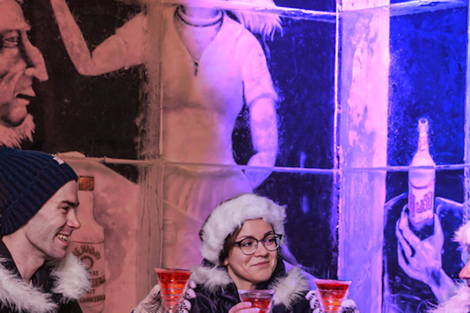 Three people in winter coats toast with drinks inside an ice bar, surrounded by ice walls with painted murals.
