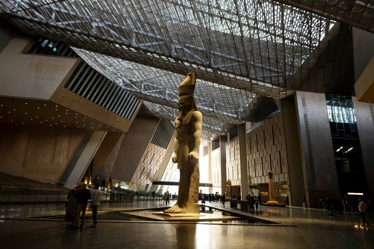 Большой крытый зал с гигантской статуей древнеегипетского фараона, современным архитектурным дизайном и небольшими группами людей.