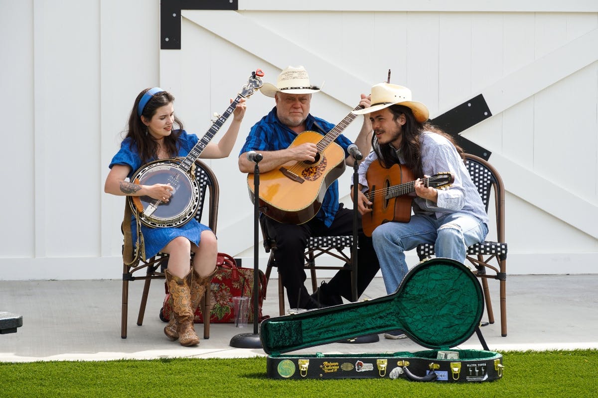 Three musicians seated, playing guitars and a banjo. One wears a blue dress and boots, the others wear hats. Guitar case on ground.