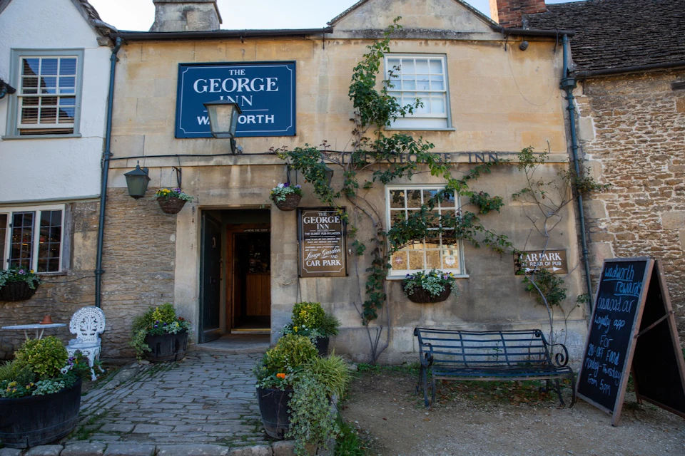 A traditional stone building labeled "The George Inn" with ivy and flowers, a bench, and a sign for parking in the front.