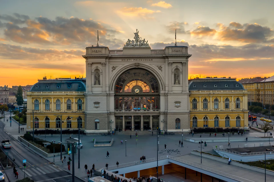 A grand, historic train station with arched windows and statues at sunset, surrounded by a plaza with people walking.