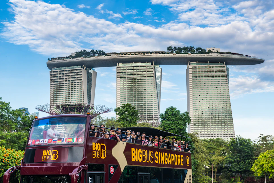 A red double-decker sightseeing bus on the street near a colorful, ornate temple structure under a cloudy sky.