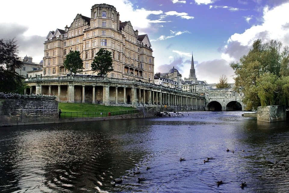 An elegant historic building with columns overlooks a river with ducks, under a partly cloudy sky.