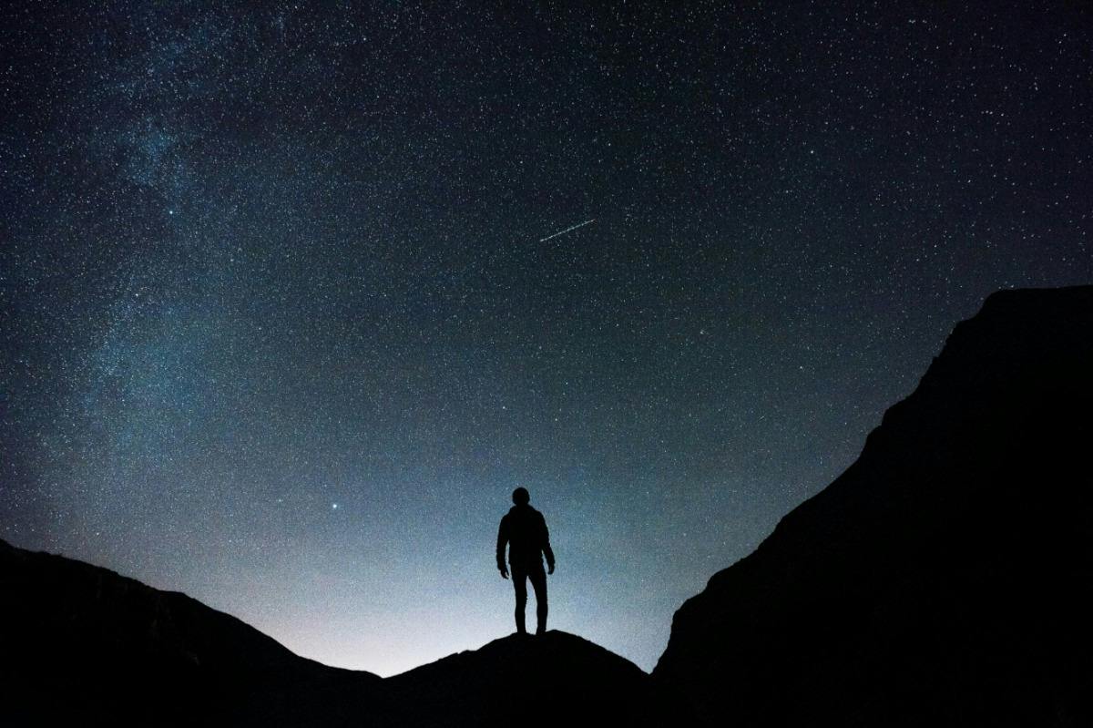A person stands on a hill under a starry night sky with silhouettes of mountains in the foreground.