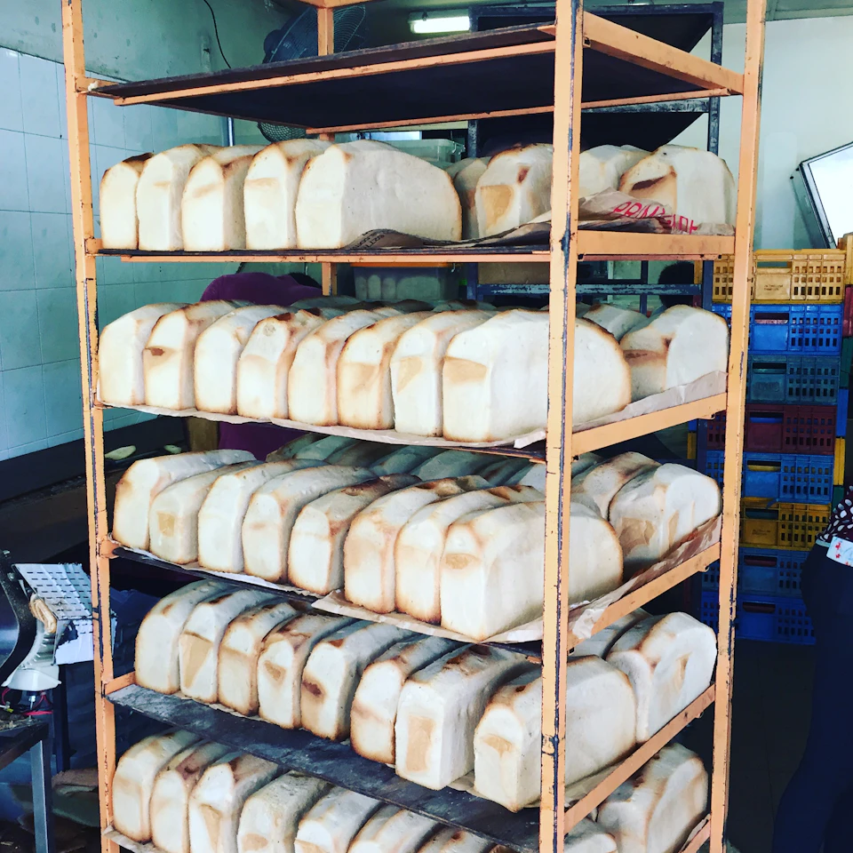 Multiple shelves filled with freshly baked bread loaves in a bakery. Blue and yellow plastic crates in the background.