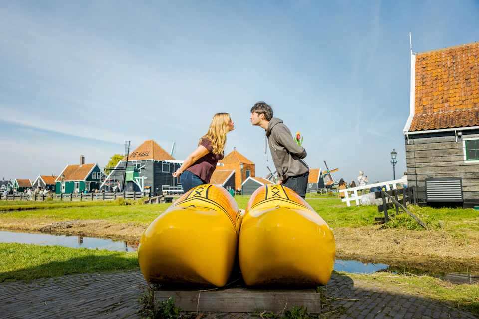 Two people lean in to kiss over large yellow wooden clogs in a grassy area with traditional Dutch houses and windmills in the background.