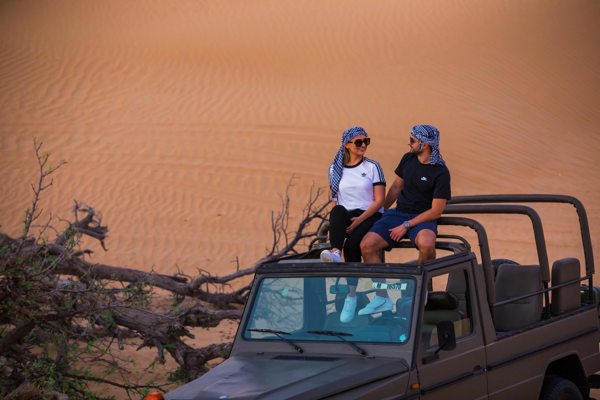 Two people, wearing headscarves, sit on the roof of a jeep in a desert setting with sand dunes in the background.