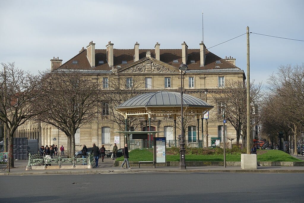 Un bâtiment historique avec un toit mansardé et un fronton orné, bordé d'arbres sans feuilles et d'un petit pavillon. Des personnes marchent sur le trottoir.