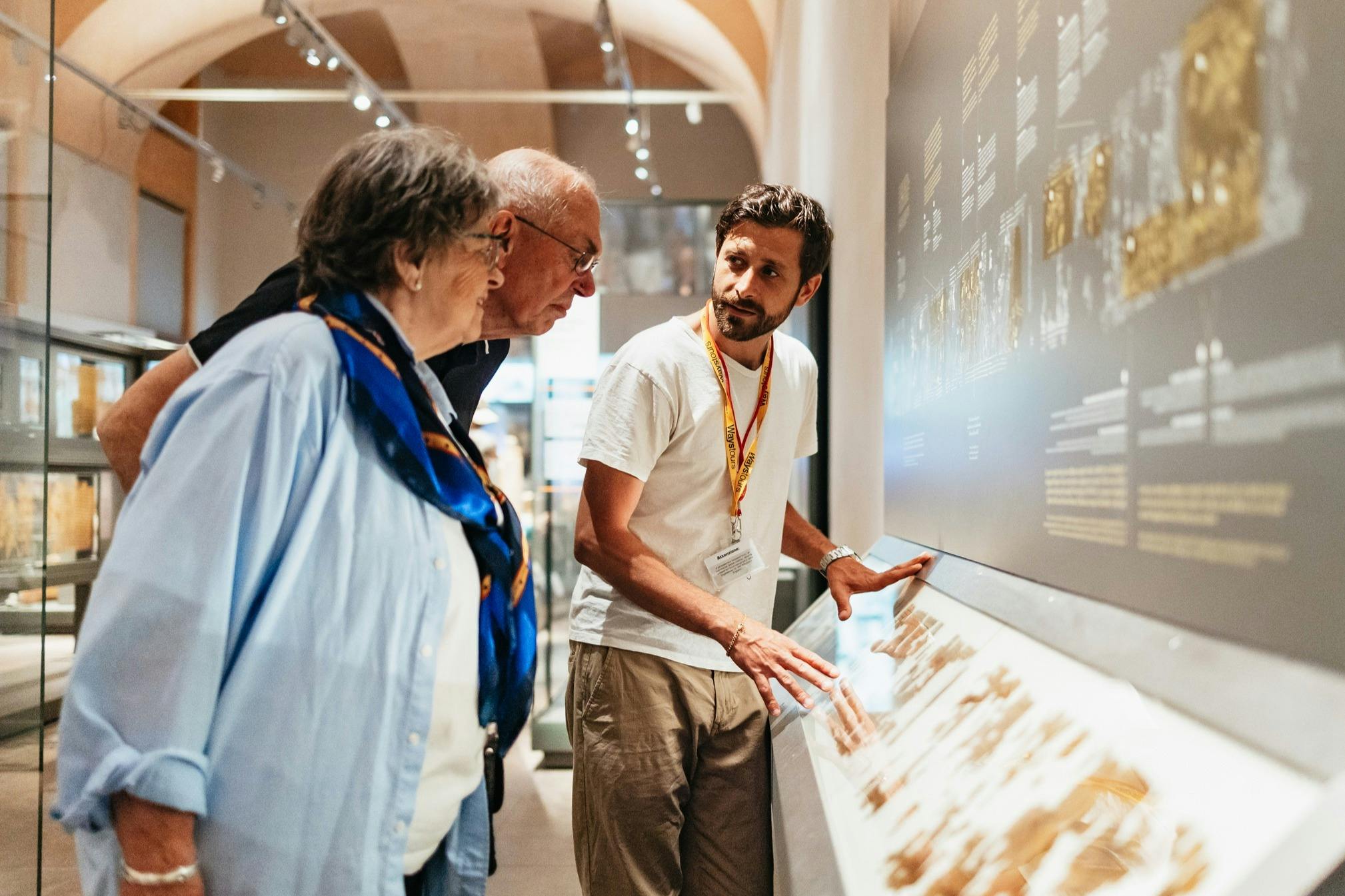 A museum guide explains an exhibit to two visitors in a well-lit gallery with a detailed display on a wall.