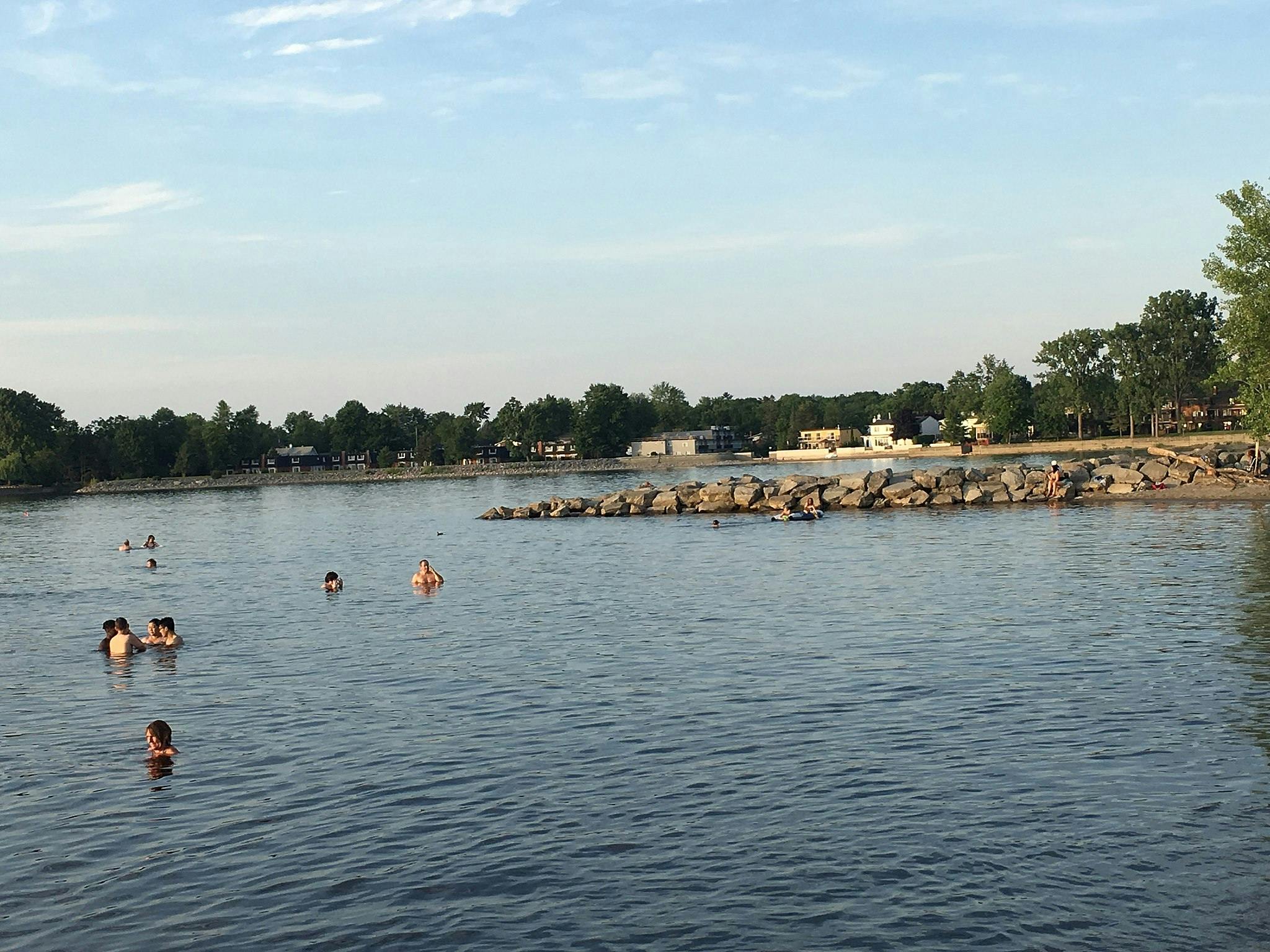 People swimming and wading in a lake near a rocky shoreline with trees and buildings in the background.