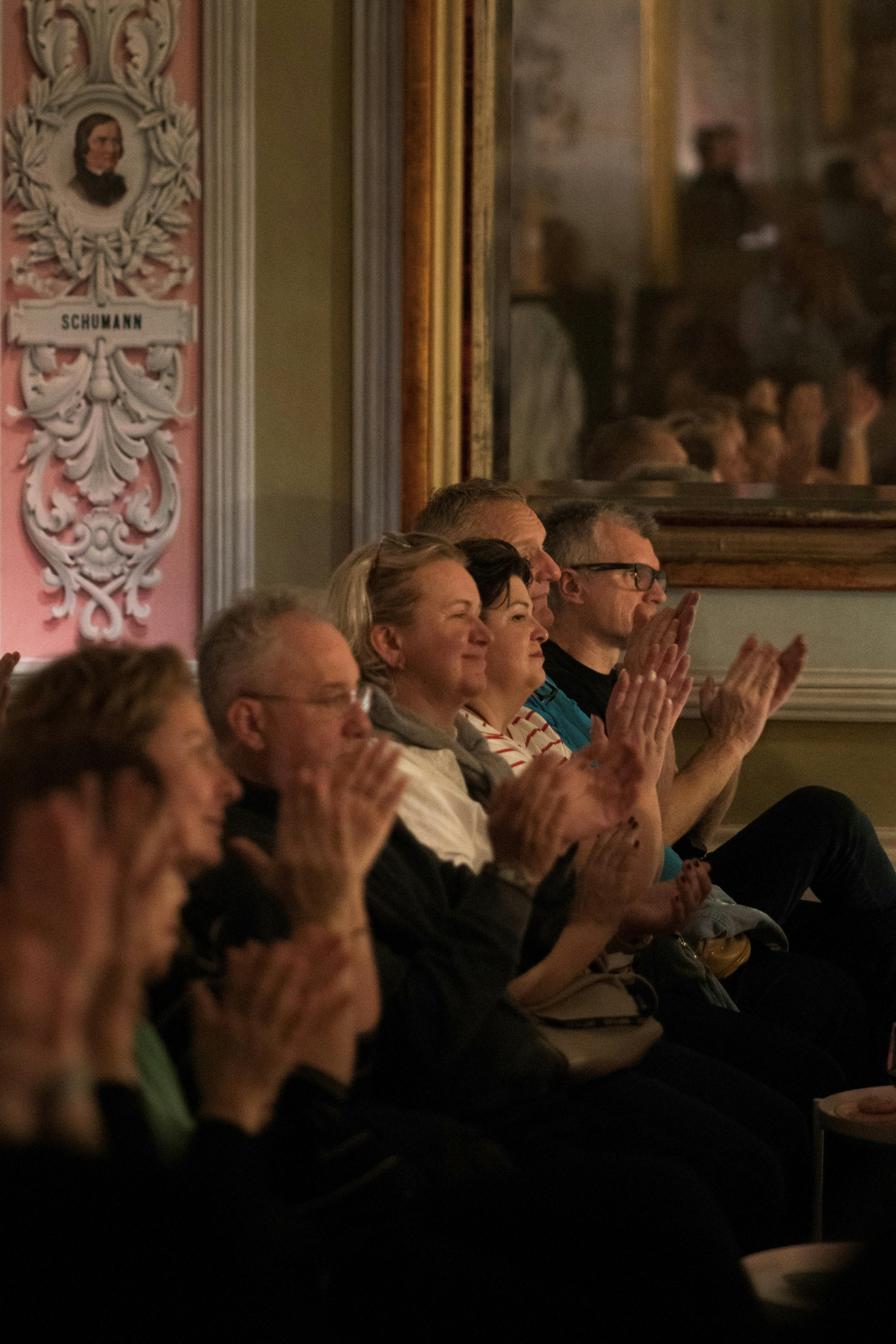 People seated in a theater, clapping. Ornate wall decoration and a sign reading "SCHUMANN" are visible in the background.