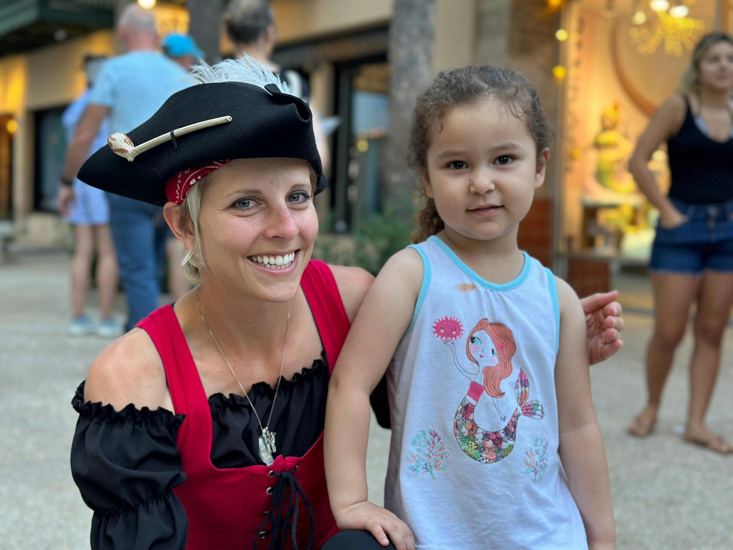Žena v pirátském kostýmu se usmívá vedle mladé dívky v košili s motivem mořské panny. Jsou venku v blízkosti obchodu.