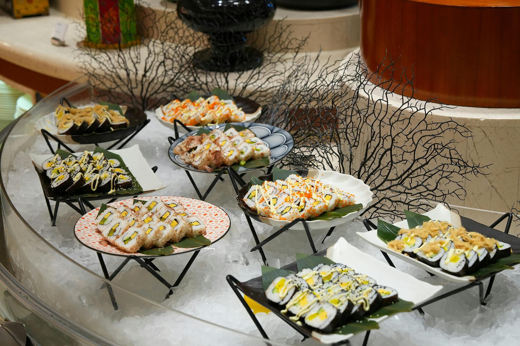 A variety of sushi rolls displayed on decorative plates over ice with intricate arrangements in a buffet setup.