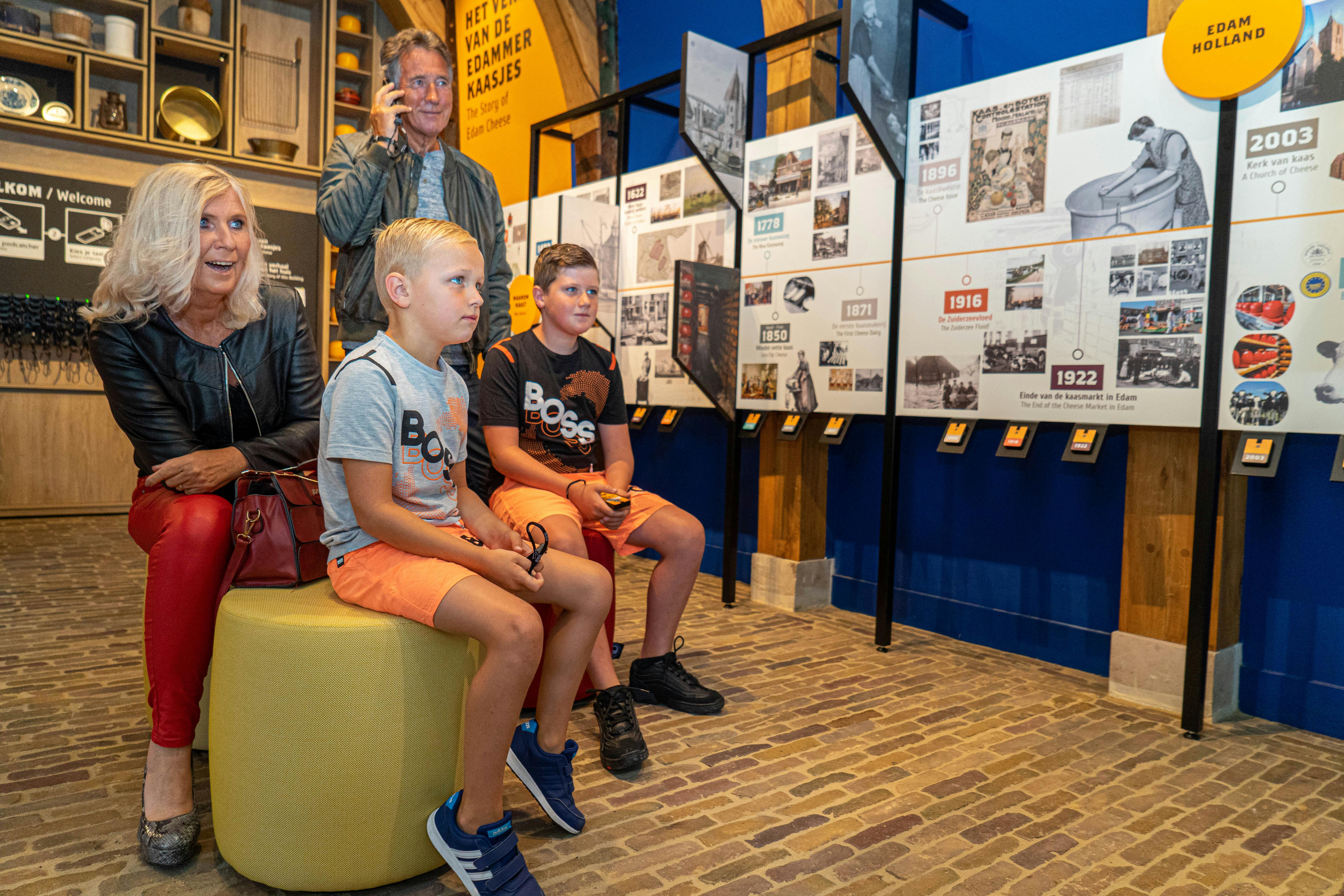 Two boys sit on a yellow ottoman, watching an exhibit with a woman and a man on a phone standing behind them in a museum.
