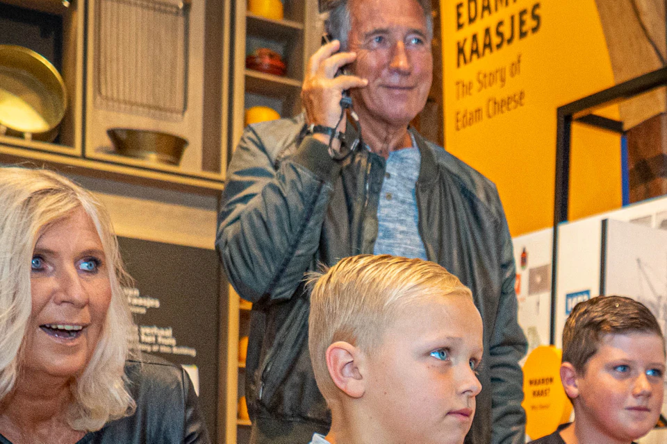 Two boys sit on a yellow ottoman, watching an exhibit with a woman and a man on a phone standing behind them in a museum.