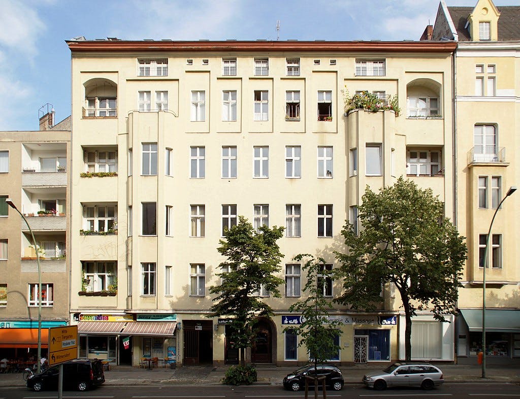 Five-story beige building with balconies, various shops on the ground floor, and trees along the sidewalk.