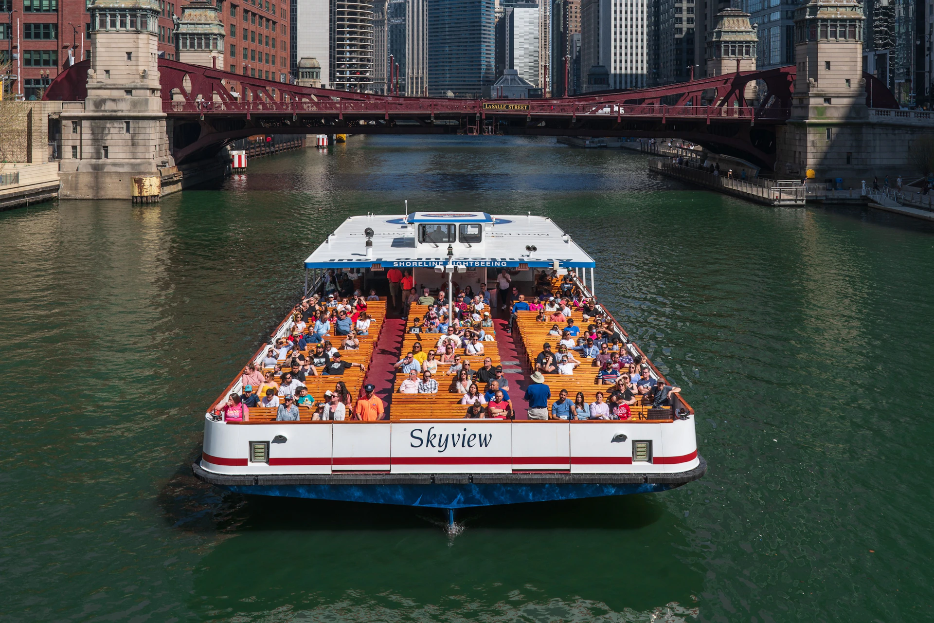 A tour boat named Skyview filled with seated passengers cruising on a river under a red bridge in a cityscape.