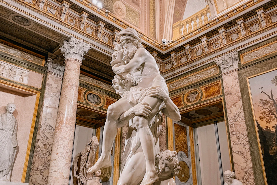 A detailed marble sculpture depicting multiple human figures in an ornate room with columns and decorative walls.