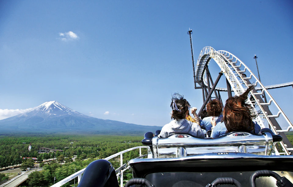 People riding a rollercoaster on a sunny day with a mountain and expansive green forest in the background.