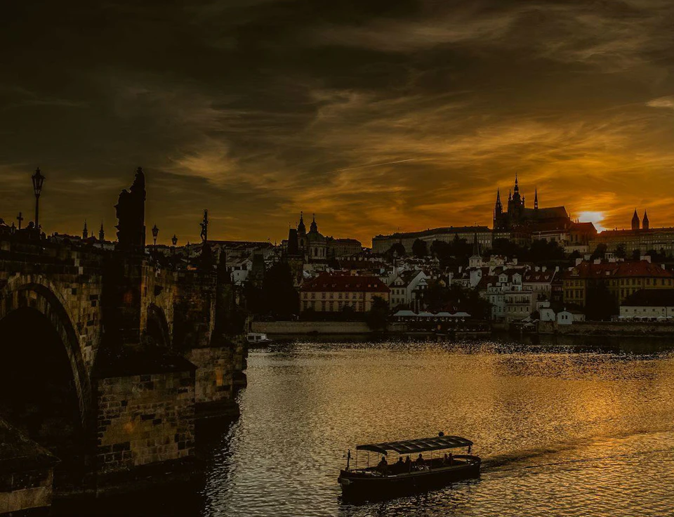 Sunset over a river with a boat, historic buildings, and a bridge in the foreground, under a dramatic sky.