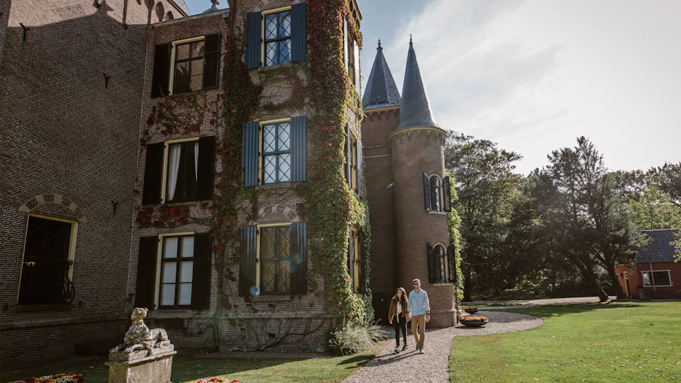 Two people walking along a gravel path beside a historic brick building with ivy-covered walls and turrets.