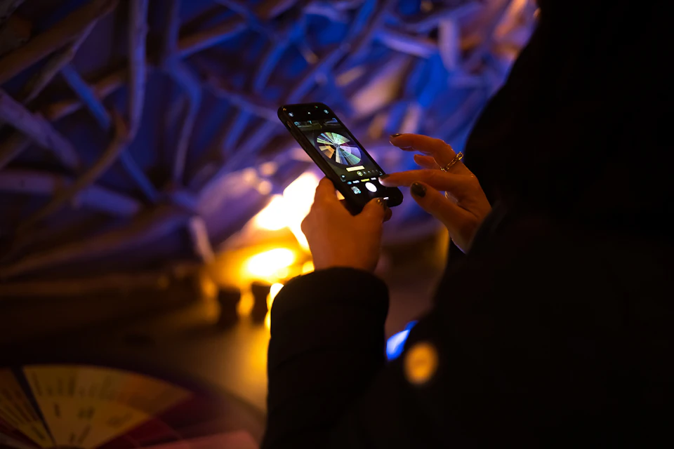 A person taking a photo using a smartphone, with a colorful pattern on the phone screen and a lit background.